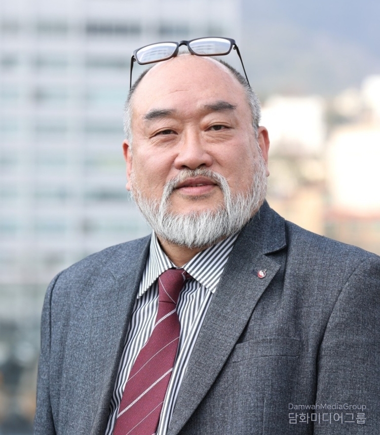  진중한 눈빛과 온화한 미소로 포즈를 취한 김용모 화백. 머리 위에 올려둔 안경은 그의 트레이드마크처럼 익숙하면서도 인간적인 면모를 드러낸다. 도시를 배경으로 서 있는 그의 모습은 예술가이자 문화행정가로서의 품격을 고스란히 담고 있다.(사진-담화미디어그룹)