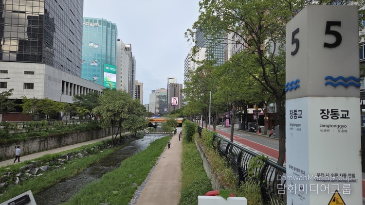 사진은 서울 도심 속 힐링 공간인 청계천 장통교 부근 전경을 담고 있다. 화면 오른쪽에는 ‘장통교(Jangtonggyo)’ 안내 기둥(5번 표식)이 자리해 위치를 알려주고 있으며, 5번 출구를 나오면 곧장 보신각 방향 도심 거리가 이어진다. 청계천 물길은 왼쪽으로 흐르고, 산책로를 따라 시민들이 여유롭게 걷는 모습이 포착되었다. 양옆으로는 고층 빌딩 숲이 들어서 있어 전통과 현대가 공존하는 서울의 풍경을 잘 보여준다. 특히 이 지점은 보신각에서 불과 60미터 거리에 있어 접근성이 뛰어나며, 청계천 산책과 종각 일대의 도심 문화를 함께 즐길 수 있는 최적의 장소임을 보여주고 있다.